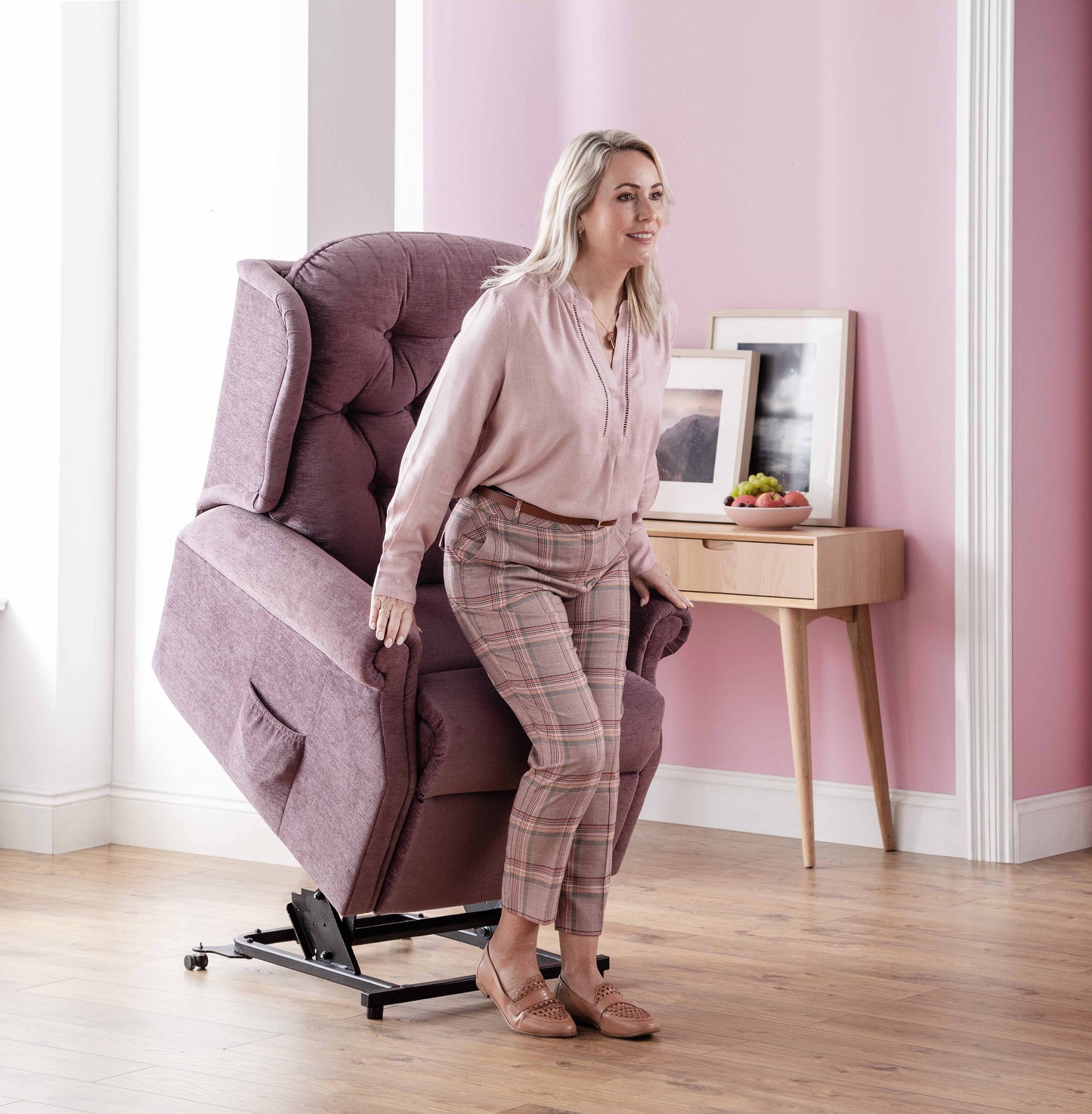 Woman using a pink Celebrity Woburn lift chair in a home setting with a pink wall and wooden side table.