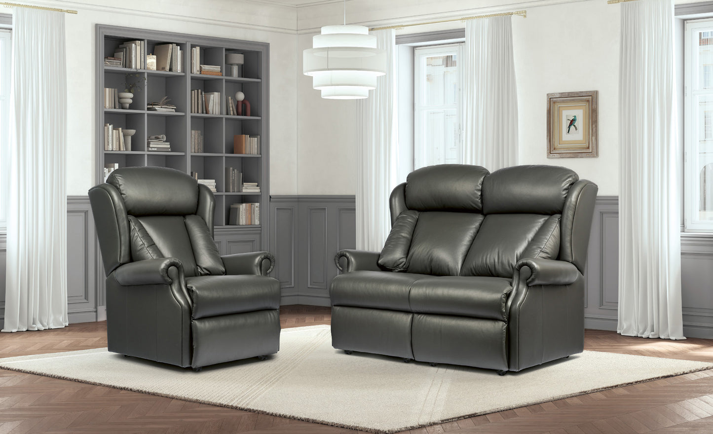 A dark gray leather recliner chair and matching loveseat arranged on a light rug in a staged living room with wood floors, white curtains, a bookshelf, and a hanging light fixture.