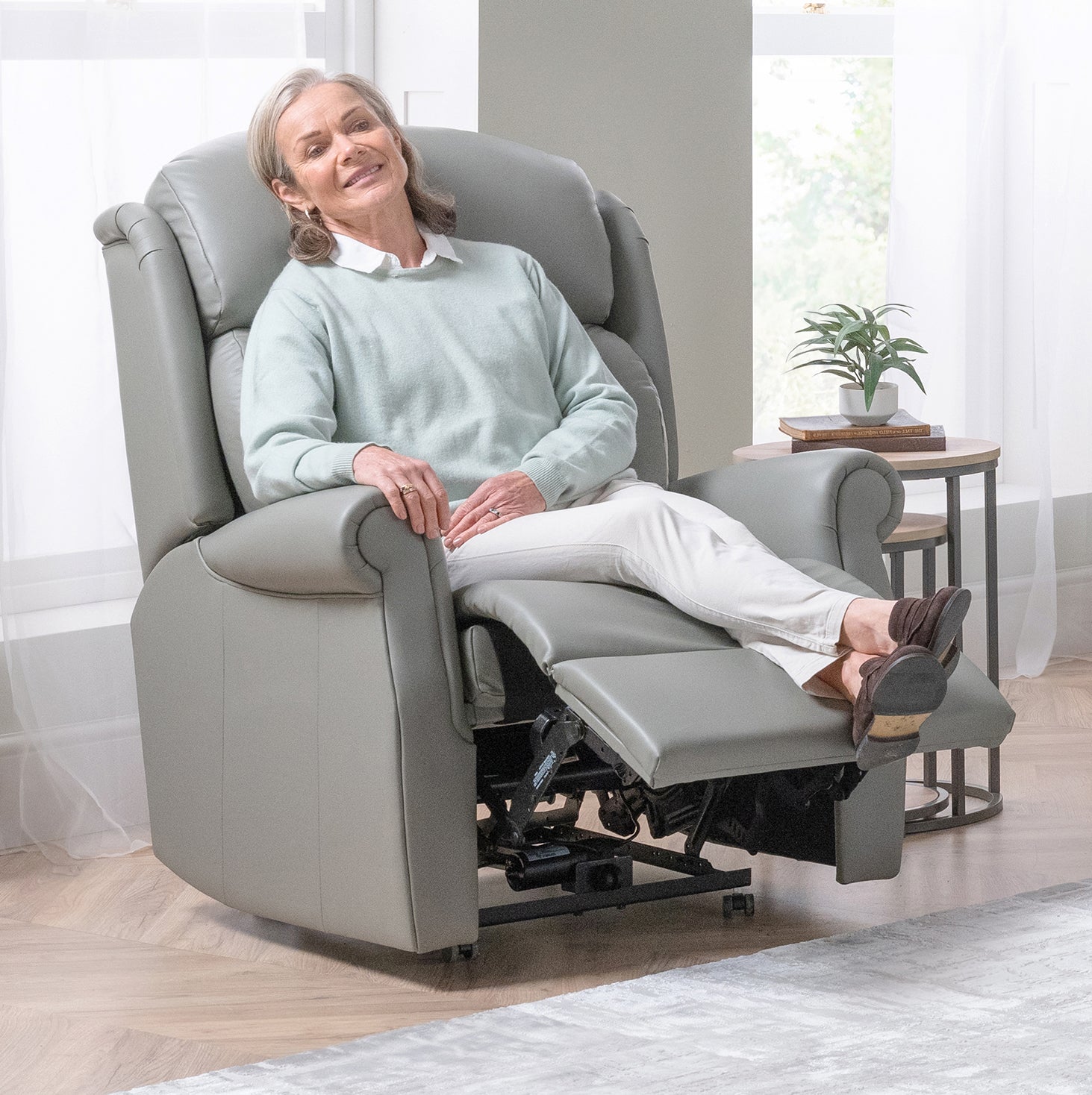 Woman sitting in a gray recliner chair in a bright room with a plant and table.
