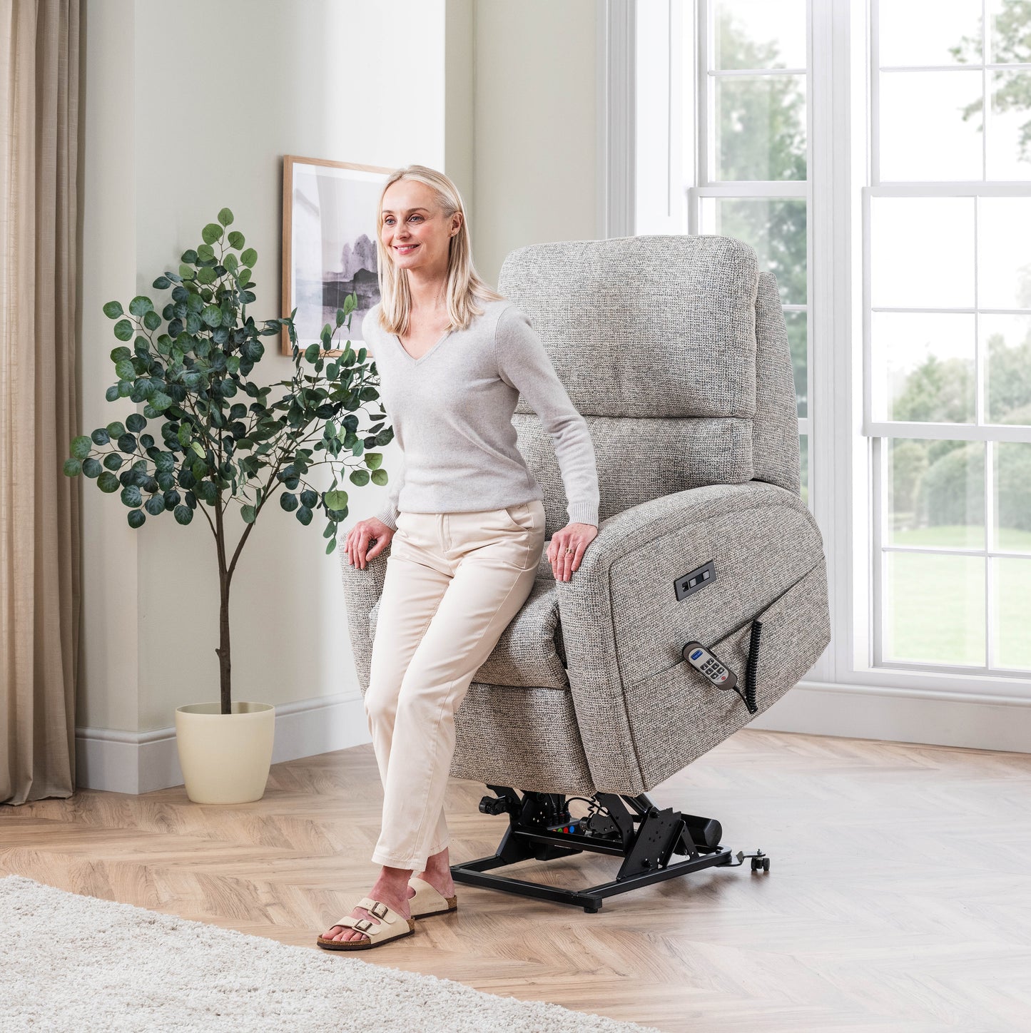 Woman sitting on a grey Celebrity Southwell recliner chair in a bright room with large windows.