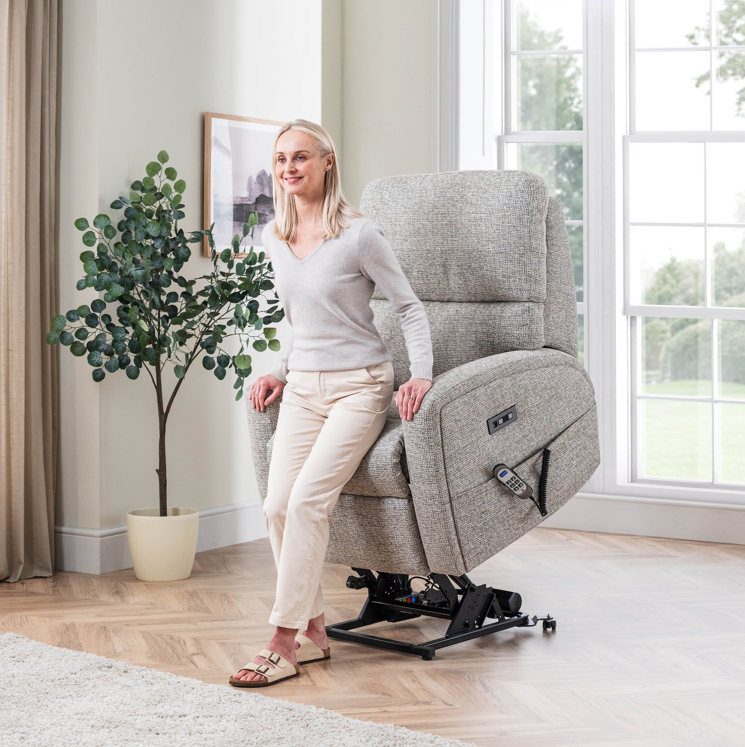 Woman sitting on a grey Celebrity Southwell recliner chair in a bright room with large windows.