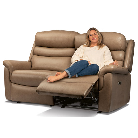 A woman reclining on a brown power lift loveseat with the footrest extended, photographed against a clean white background.